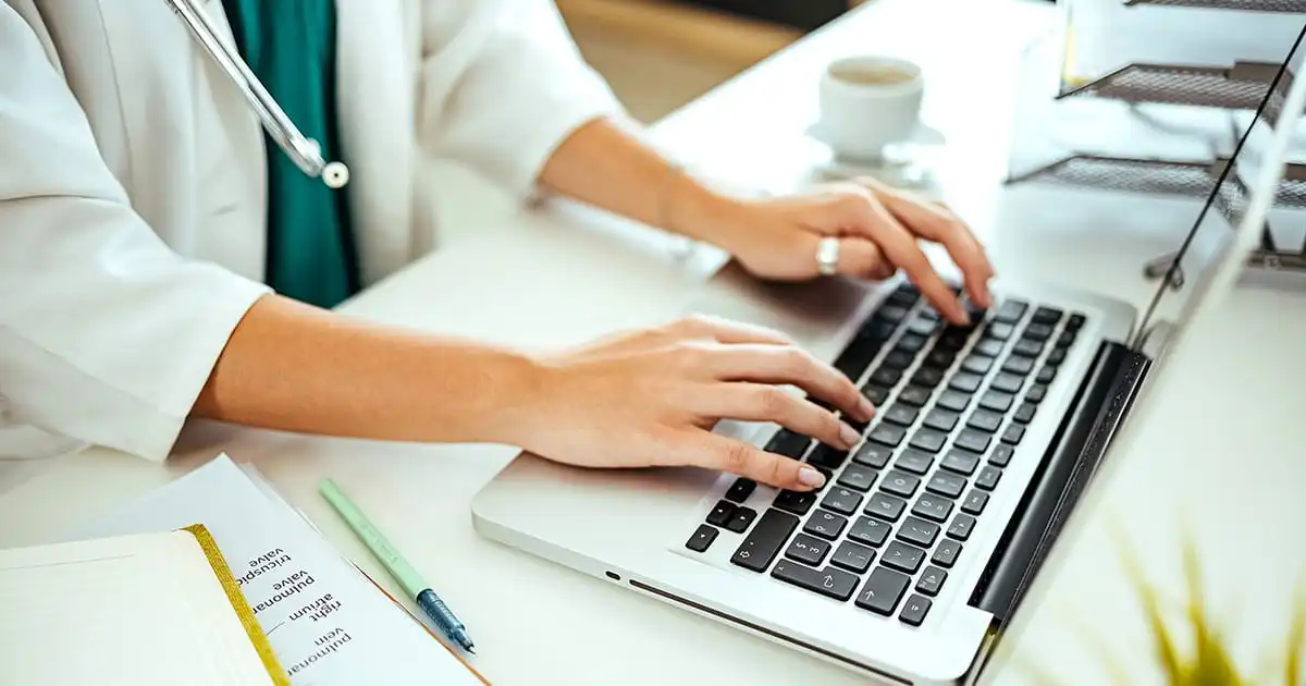 A person in a white coat types on a laptop at a desk with a clipboard, pen, and coffee cup nearby, representing the professionalism required for Provider Credentialing & Enrollment Services in USA