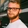 Bearded man with glasses and white shirt stands outdoors in front of a building and bus.