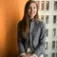 Woman with long brown hair in a gray blazer sits by a window holding a notebook; office setting with orange wall.