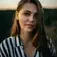 Woman with long brown hair in a striped shirt smiles outdoors at sunset, greenery blurred in the background.